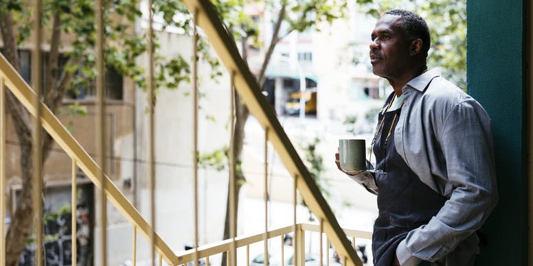 Side view of a Black man in an apron, standing on outdoor stairs with cup of coffee and looking away from the camera.