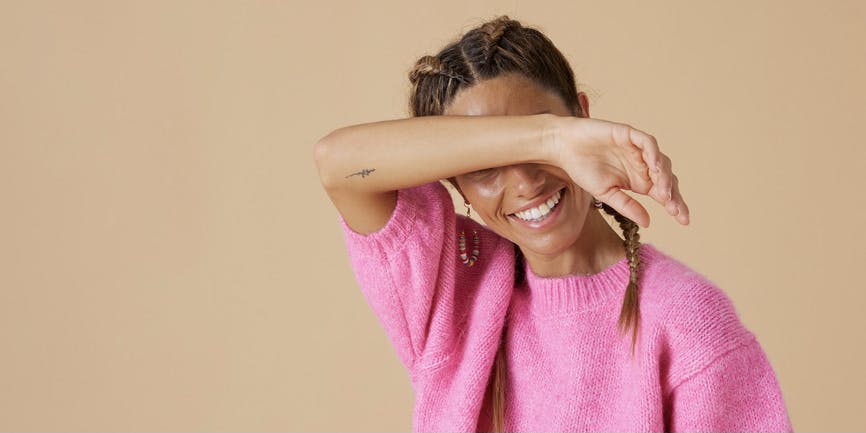 Joven mujer negra riendo con las manos cubriéndose los ojos con un suéter rosa casual