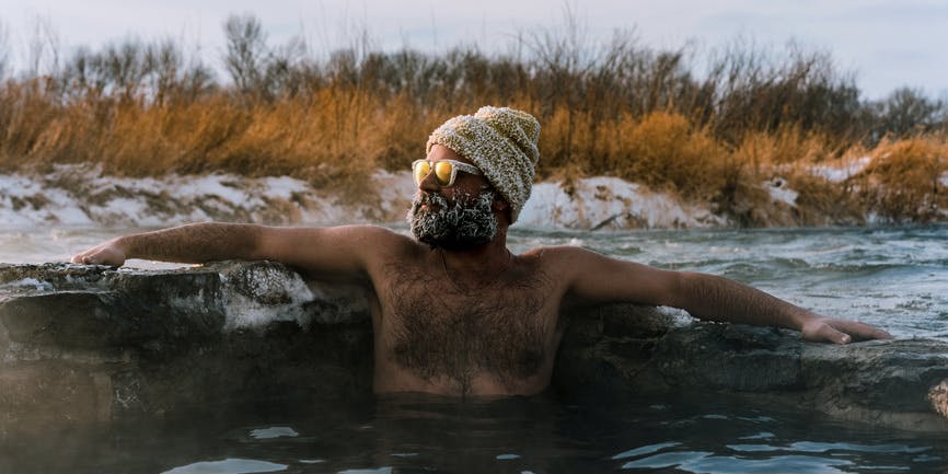 Brown-skinned man relaxes in a Renova Hot Spring, wearing a towel wrapped around his head and glasses, with his beard frozen and a snowy, brushed landscape behind him.