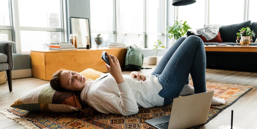 A woman in casual clothes smiles while using a mobile phone lying on the floor at home