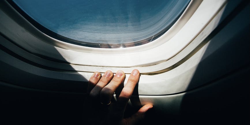 A color photograph of a hand in shadow pressing fingers against an airplane window with a blue sky and sunlight streaming in.