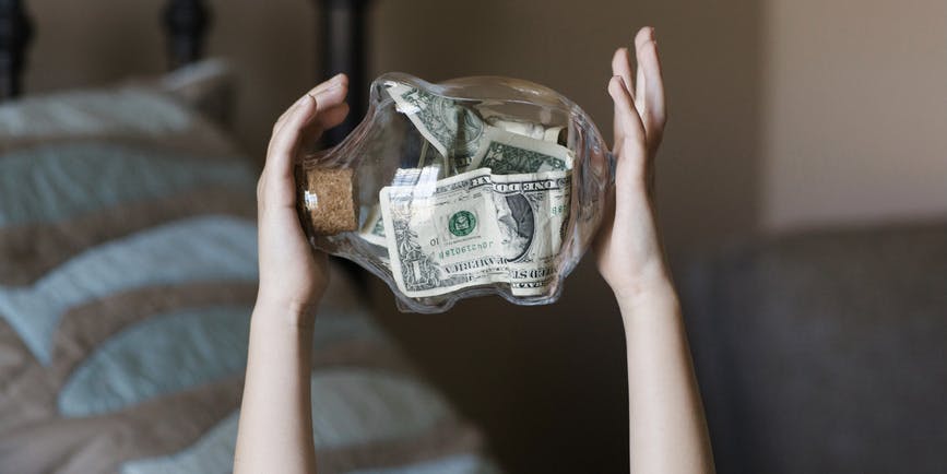 A child's hands old up a clear piggy bank with a few dollars in at against a blurry background of a bed and chair.