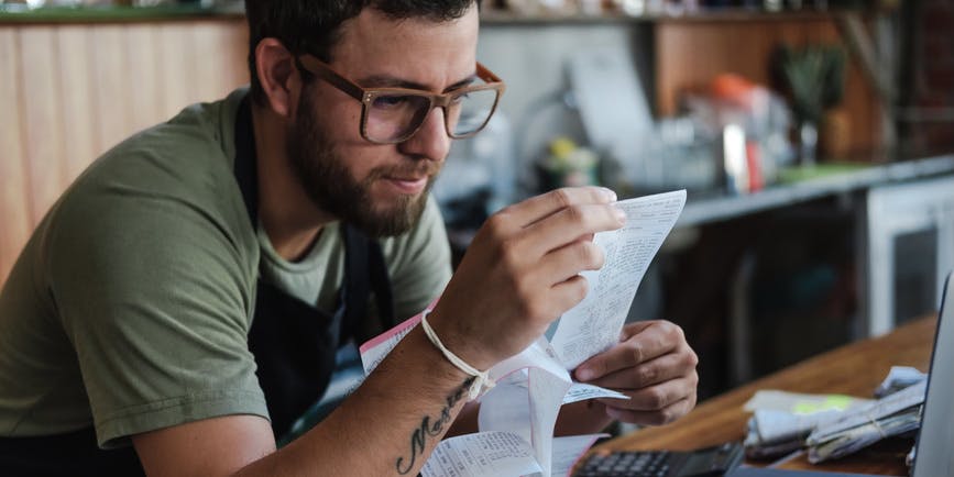 A young Latino man wearing glasses and a green t-shirt works at the counter of his business, looking intently at a receipt while working on finances. 