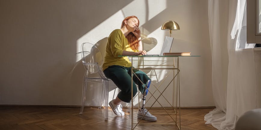 An attractive white disabled woman with red hair and a prosthetic leg enjoys the rays of the sun in her home office.