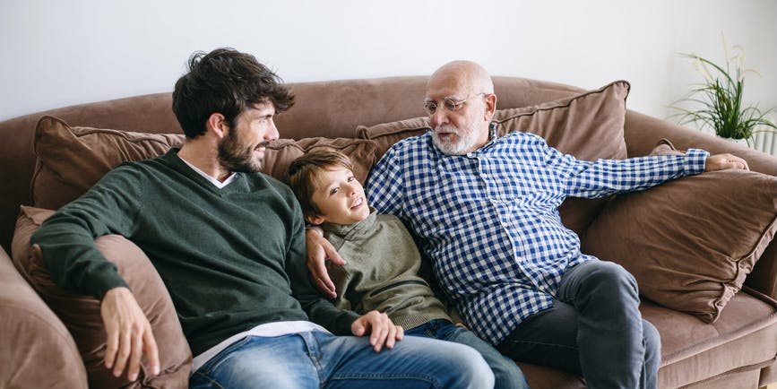 A grandfather, father and son at home on the couch, talking and smiling.