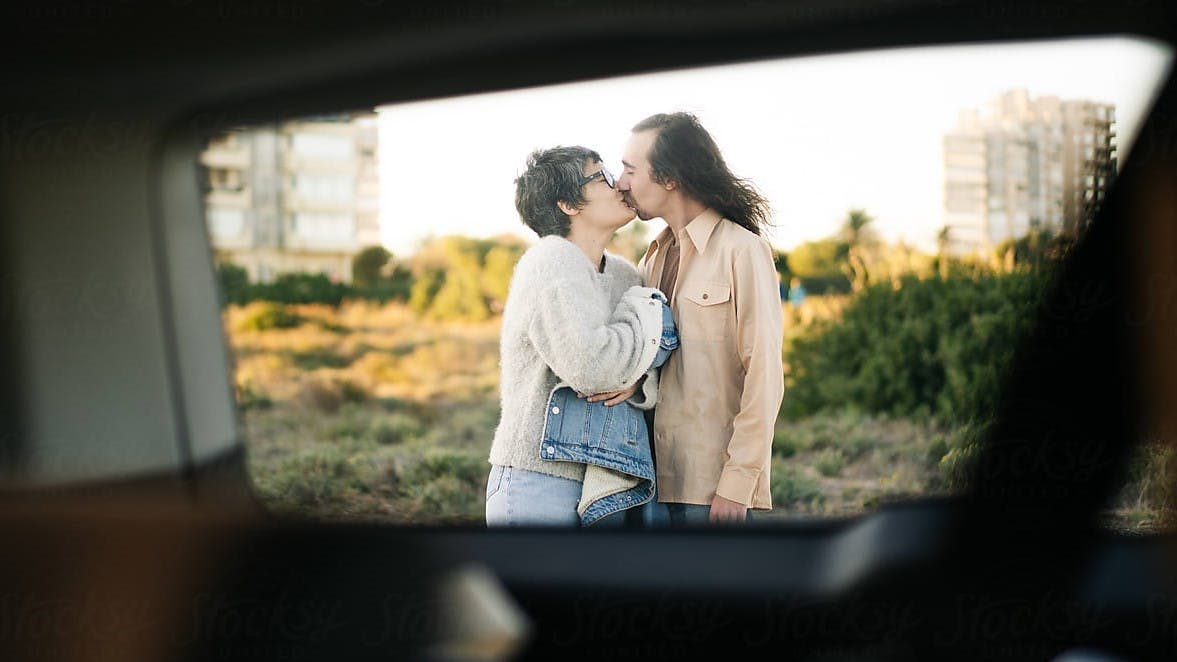 Adult man and woman in retro outfits kissing each other while standing near car in city outskirts