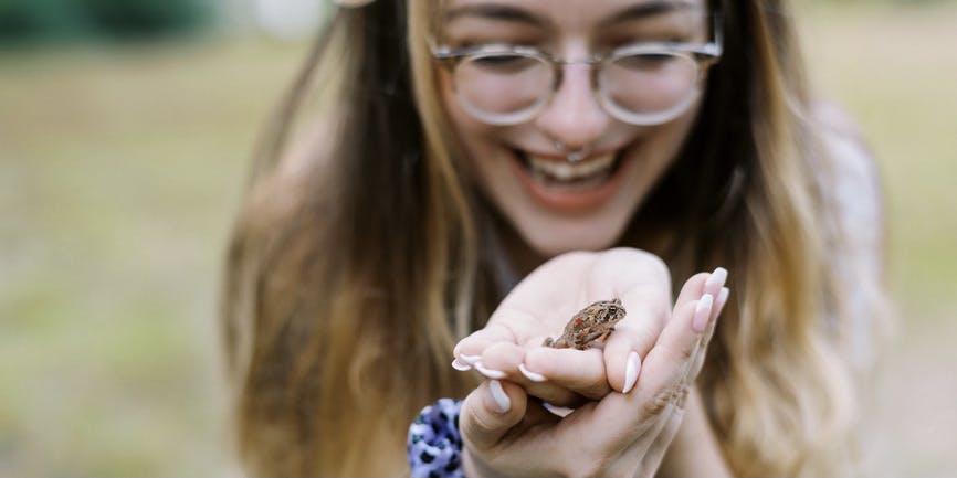 A happy white woman with long blond hair, glasses, and a pierced nose smiles as she holds a small toad in her palm.