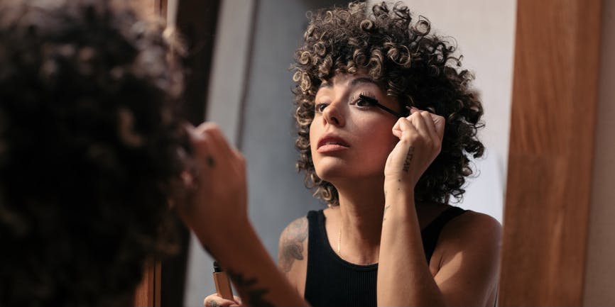 Curly haired woman with brown skin and tattoos applies mascara at home in front of a mirror.