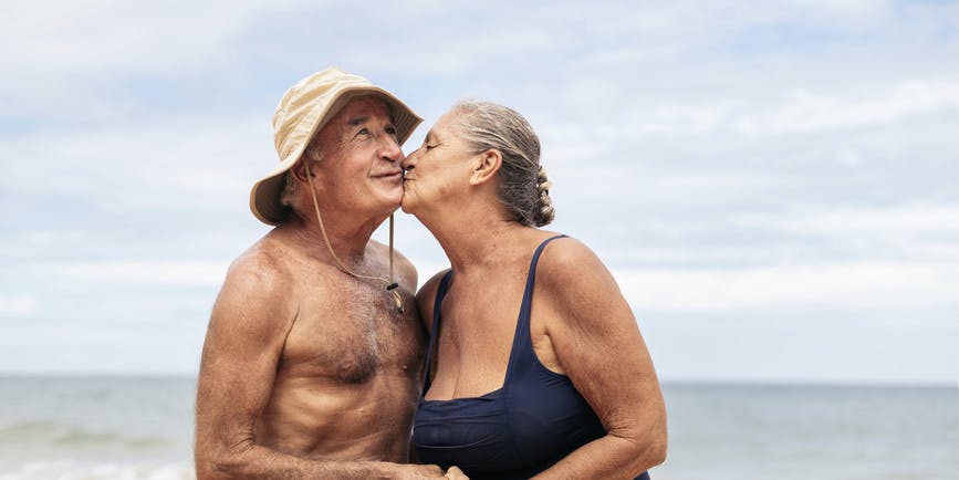 Color photograph of a senior white couple on a beach wearing bathing suits, smiling and embracing each other.