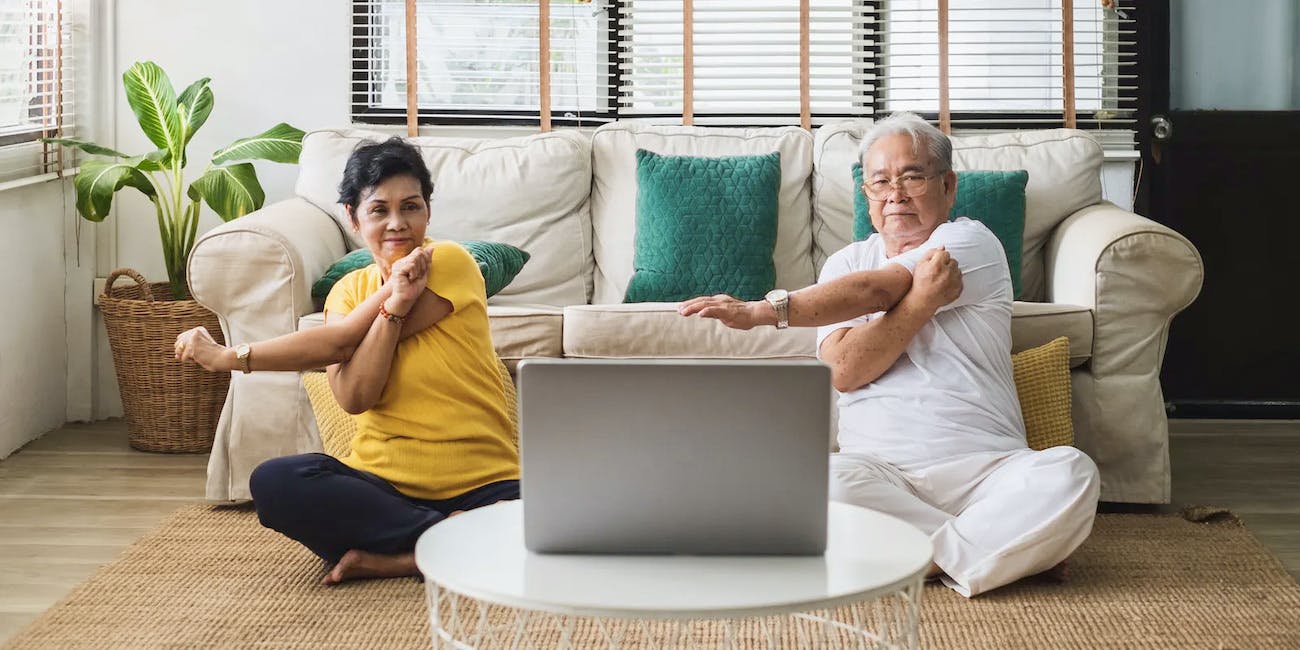 A senior Asian couple sits on their floor looking at a computer, stretching their arms while doing yoga with online instruction.