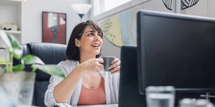 A white woman with short brown hair sits in front of a computer in a modern, colorful office holding a cup of coffee as she smiles and looks up at someone off camera.