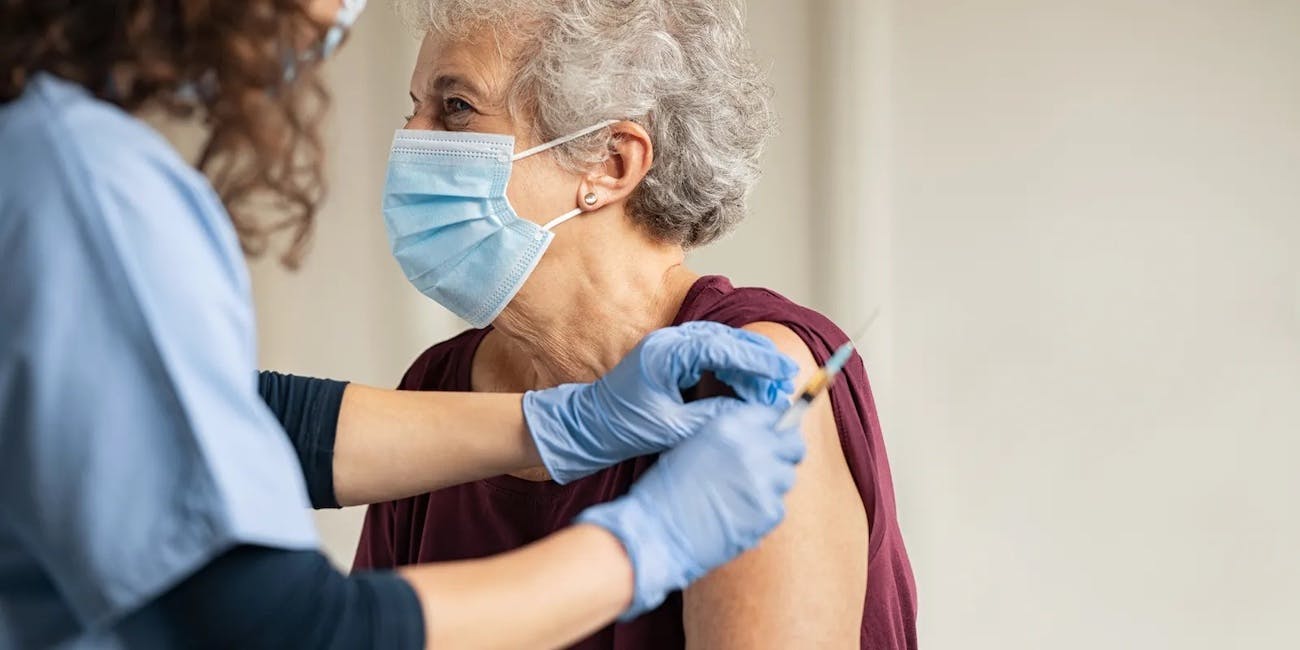 A white senior woman wearing a dark red shirt and medical mask gets a vaccine in her left arm from a practitioner wearing scrubs and a medical mask.