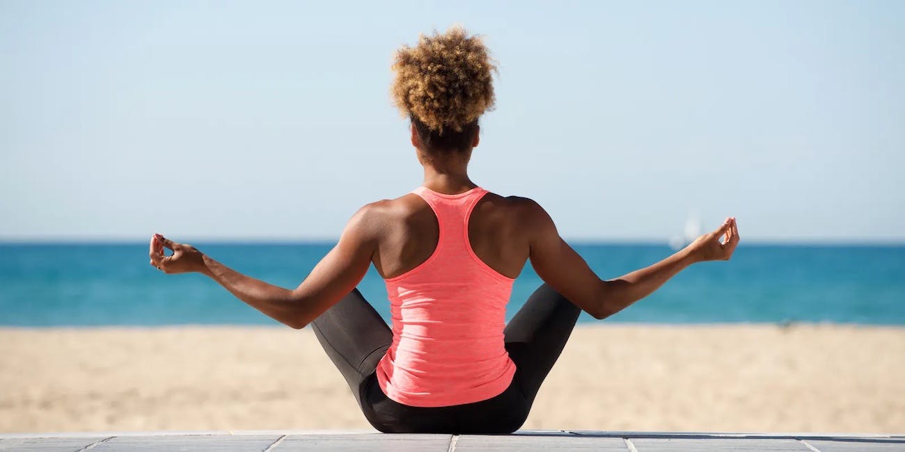 A toned young Black woman wearing black leggings and a bright pink fitted tank sits on the beach looking away from the camera as she holds an elevated yoga pose.