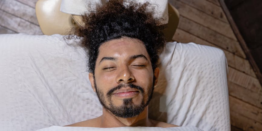A young Black man with a beard and Afro lays on a massage table with his eyes closed, smiling.
