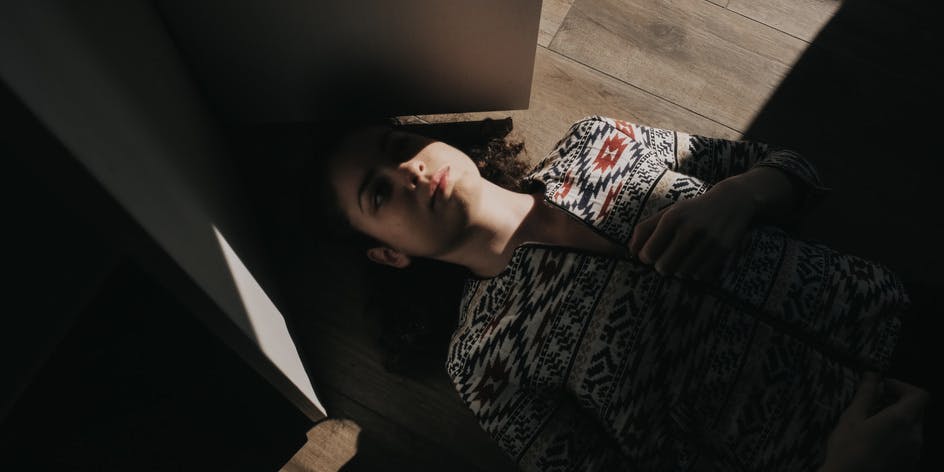 A. young white woman wearing a colorful cardigan lies on a floor with her eyes closed, half hidden from the sunlight by a half-closed door.