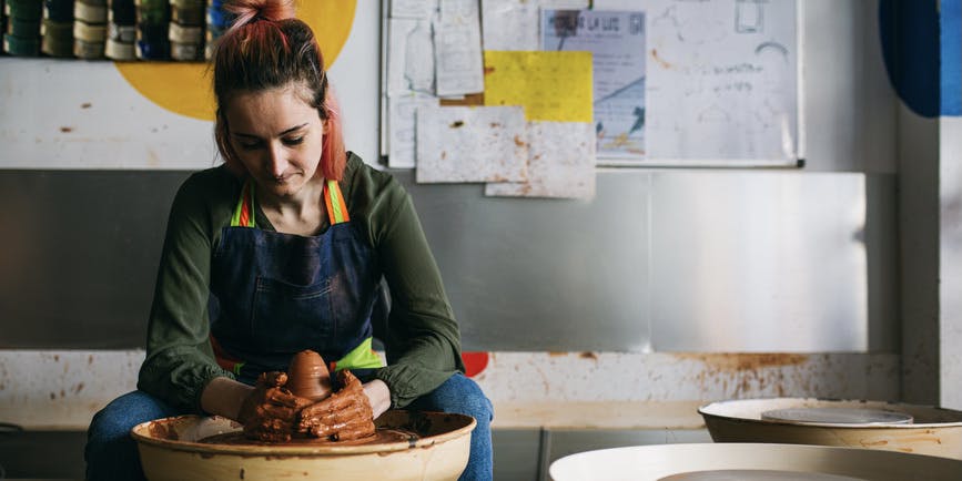 A young white woman with pink and brown hair tied back wearing a green shirt and blue apron with rainbow ties sits looking down at a potter's wheel as she works clay with her hands.