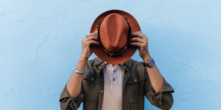 Hombre hispano irreconocible cubriéndose la cara con un sombrero sobre un fondo azul vibrante.