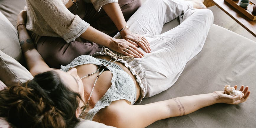 An anonymous white  woman giving an energy healing (reiki) to another white woman with menstrual pain. Both are dressed in natural fabrics and wear a mala around their necks. The woman receiving the treatment is holding a crystal in her right hand.