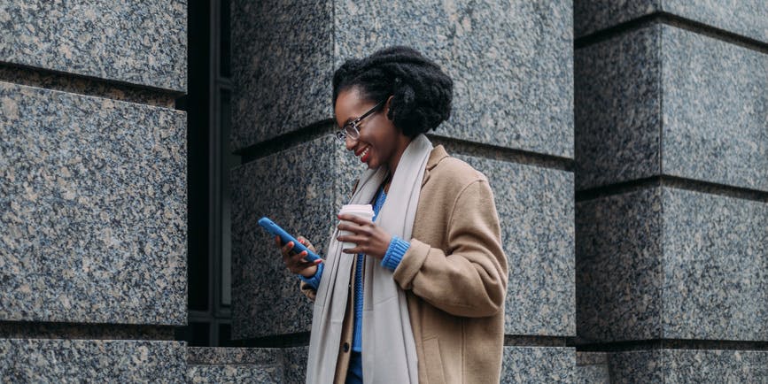 Una joven negra de pelo negro corto, vestida con un abrigo largo de lana tostada, un suéter azul y una bufanda larga color canela camina junto a un edificio de piedra gris de la ciudad, sosteniendo una taza de café para llevar y sonriendo mientras mira su teléfono.