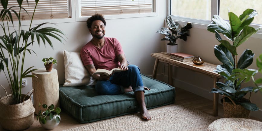 Full body photograph of a young Black man in casual clothes laughing at a joke while sitting on a soft floor pad and reading a book at home in a sunlit room surrounded by plants and wood and rope accents. 
