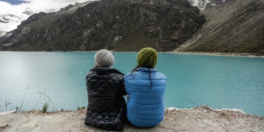 Two women seen from behind sit on the edge of a large blue lake surrounded by gray slate mountains, wearing winter coats and hats, looking out at the water. 