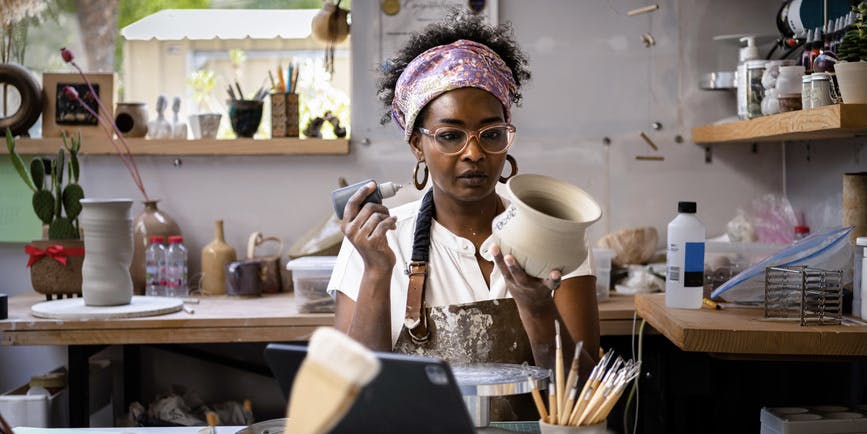 A stylish Black woman wearing a brown leather apron, glasses and violet bandana sits in a ceramics studio surrounded by instruments, concentrating on a pot in her hand.