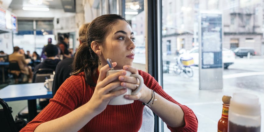 A young Latina woman wearing a red longsleeved shirt sits in a New York City diner holding a mug of coffee and looking out the window.