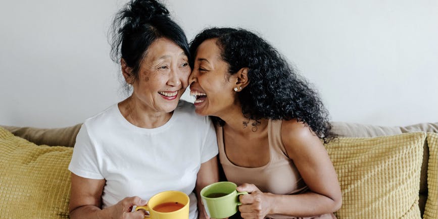 Senior asian mother with laughing adult daughter talking while drinking tea sitting on couch at home