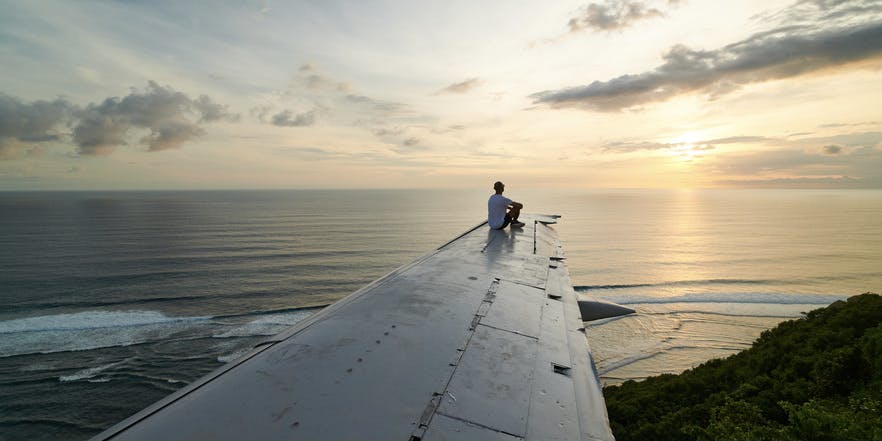 Una fotografía de ensueño de un hombre sentado en el ala de un avión cerca del océano, mirando fijamente la lejana puesta de sol.