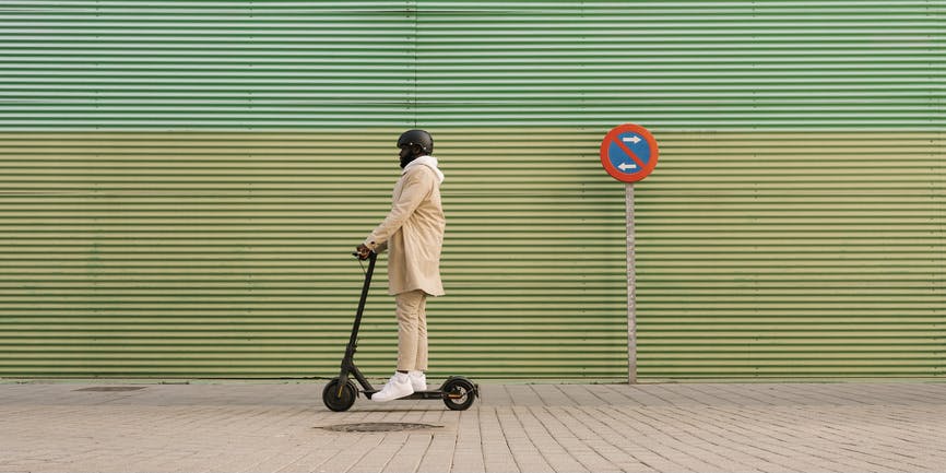 A Black man wearing a pale pink suit and helmet rides an electric scooter down a London sidewalk.