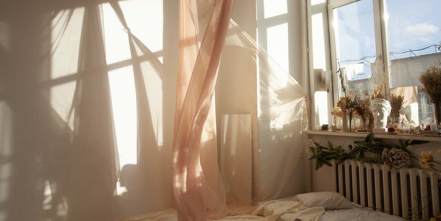 A dreamy indoor photograph of a bedroom where a gauzy curtain dances in the breeze of an open window and sunlight casts shadows on the wall.