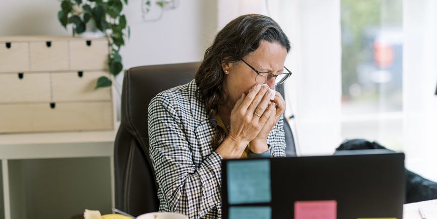 An older female professional with long graying hair wearing a checkered jacket blows her nose at her desk.