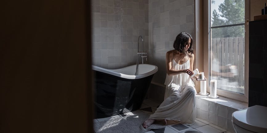  A dreamy photo of a young white woman in a long white nightgown lighting candles in a bathroom with large checkered tiles and a claw foot tub, with light streaming in from the windows.