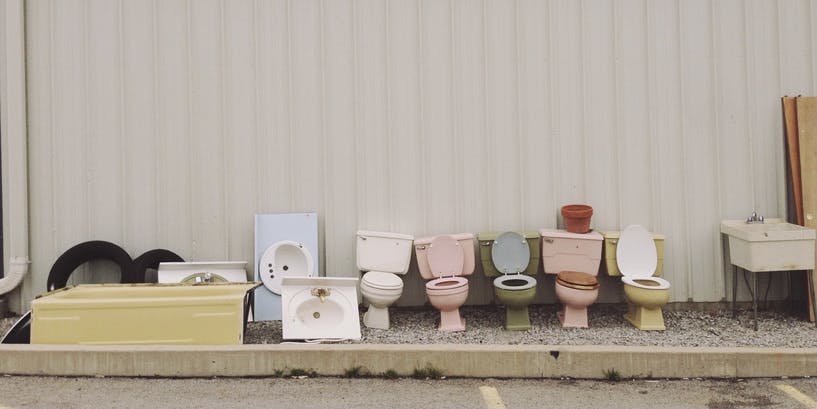 An outdoor photo of a pink exterior wall with discarded toilets in various colors lined up against it.
