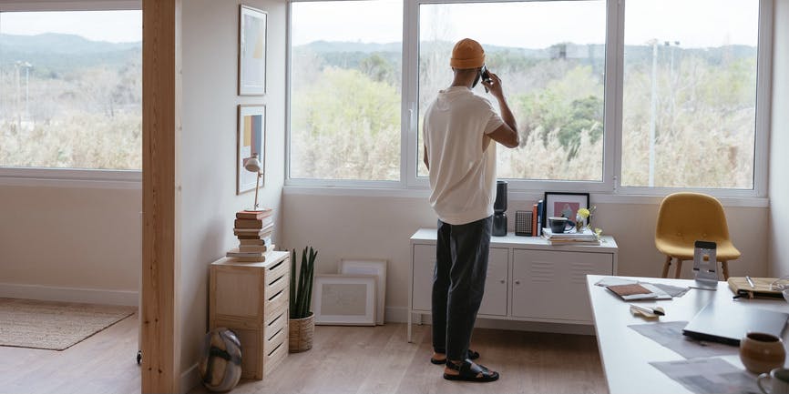 A young Black man stands in a modern office with his body looking out a window, away from the camera, talking on a cell phone while gazing out onto mountains of trees.