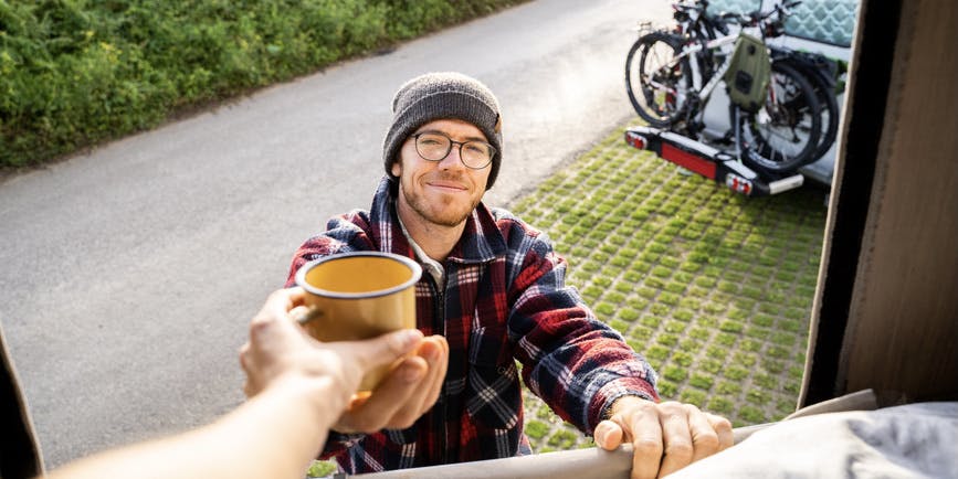 Man standing next to his camper van with bicycles in the morning giving a coffee mug to friend in van.