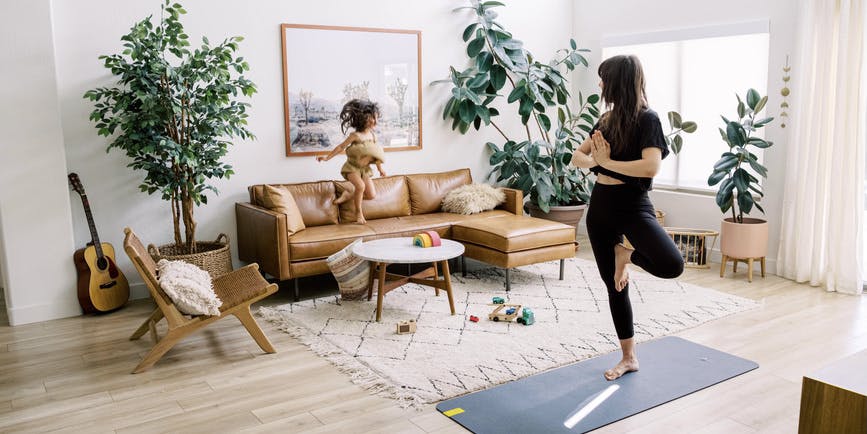 A colorful photo shows a young white mother doing yoga on a mat in her airy, bohemian living room while her young child jumps around on a leather couch.