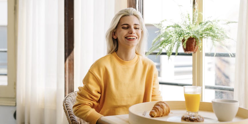 Una joven blanca de cabello blanco y un jersey amarillo sonríe mientras está sentada en un café bien iluminado con un croissant y un vaso de jugo sobre la mesa frente a ella.