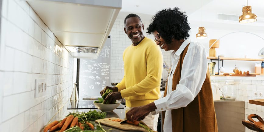 Cheerful Black man and woman in trendy casual clothes smiling and looking at each other while standing near table with fresh vegetables and cutting vegetables for salad in kitchen.