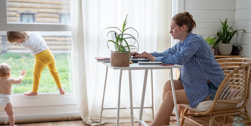 A young white mother wearing a long sleeved shirt and no pants sits slumped in front of a computer at a small table while her toddler and baby play by the windows of her airy, modern home.