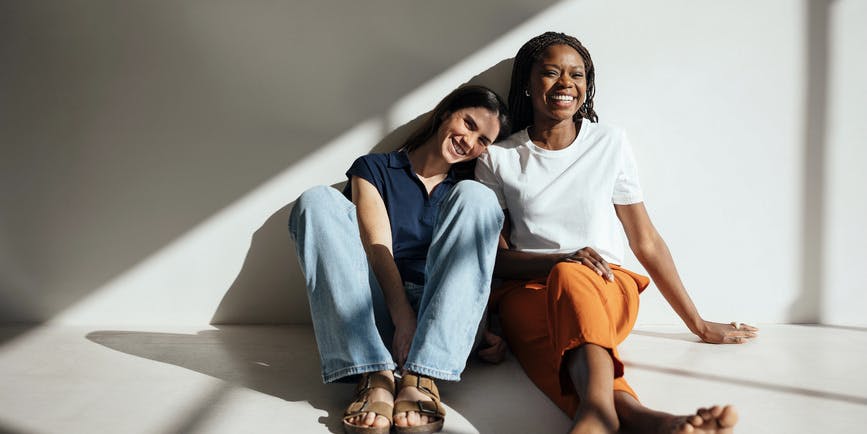 Portrait of happy women in trendy clothes spending time together while sitting on floor in bright studio