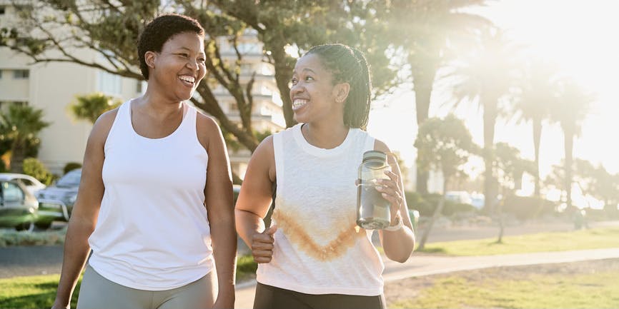 Two young Black women walking outdoors exercising and working out in an urban park. They are happy and wearing workout clothing and carrying a water bottle.