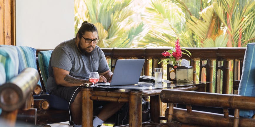 Shot of a young brown man wearing glasses working online on his laptop outside in a tropical climate while having a drink.