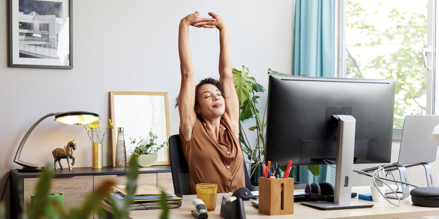 Joven oficinista con los brazos levantados y los ojos cerrados sentada en el escritorio con el ordenador mientras trabaja en la oficina
