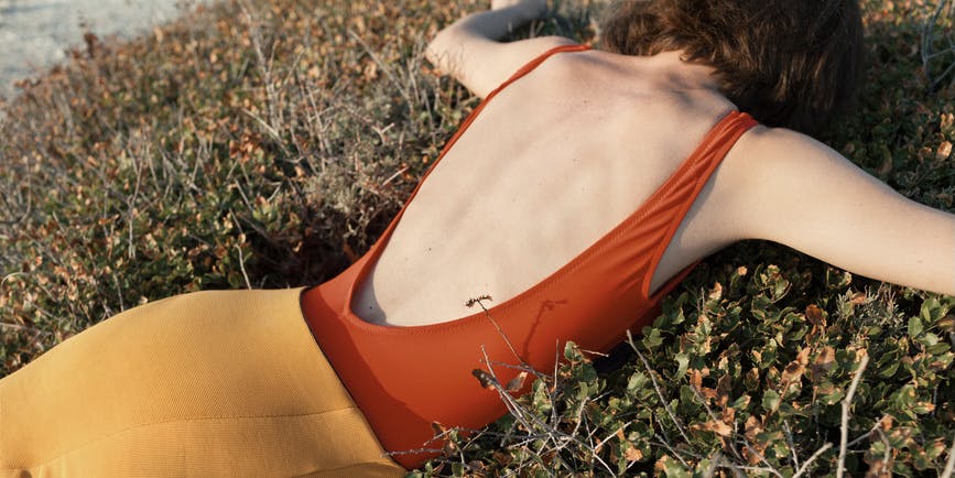 A white woman with short red hair wearing a dark red strapless body suit and orange skirt lies facedown in a bush.