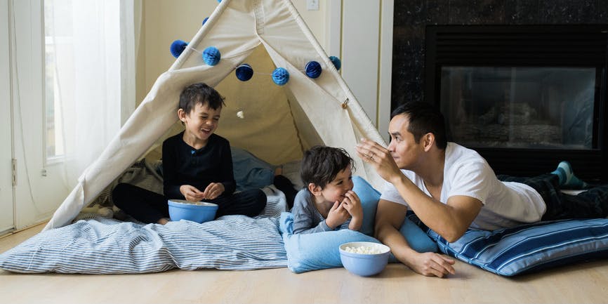 A young Latino father plays on the floor with his two sounds, underneath a tent in their living room, laying on top of blue pillows, snacking from bowls of popcorn.
