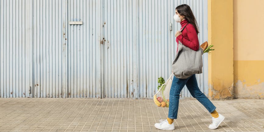 A young thin woman with long brown hair wearing jeans, a sweater and a face mask walks on a city street returning from shopping, with fresh fruit, vegetables and bread in reusable bags.