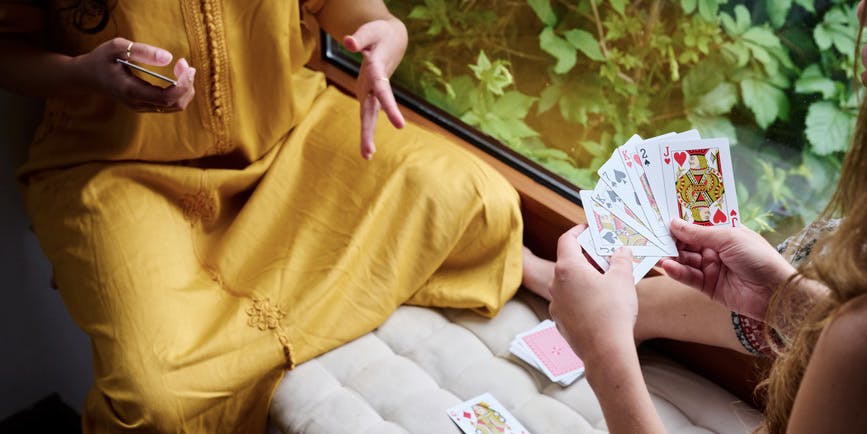 Two unrecognizable cropped white girlfriends in spring clothes playing simple card game in a window seat with greenery outside. One explains the rules, the other girl holds a spread of cards in her hand.