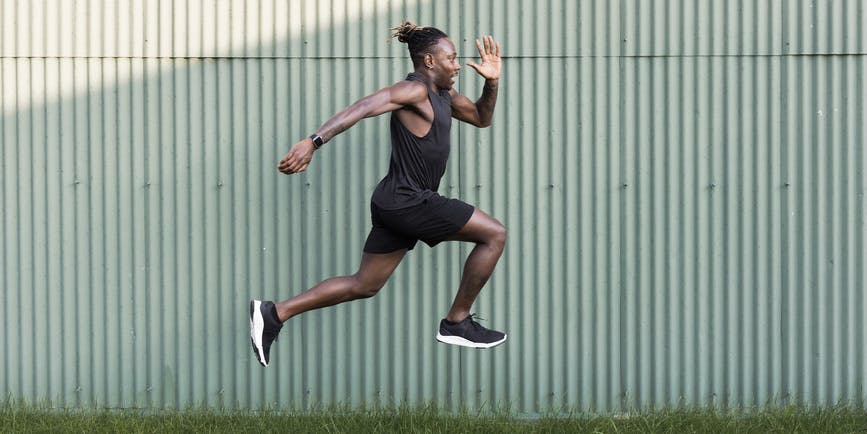 Young Black man wearing workout clothes doing physical exercises by taking big jumps outdoors against a stamped metal wall with tufts of grass on the ground.
