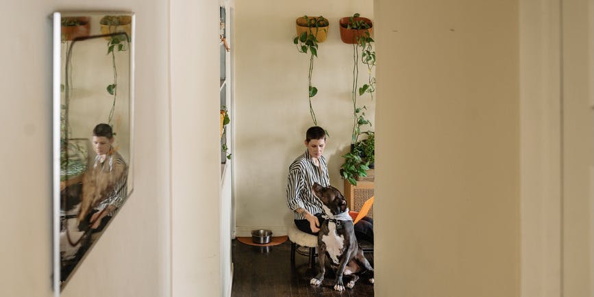 LGBTQ+ woman inside their home working on laptop while petting their large dog who is sitting on the floor next to them. The photo is taken from far away with walls and a doorway framing the shot and part of their reflection shown in a side mirror.